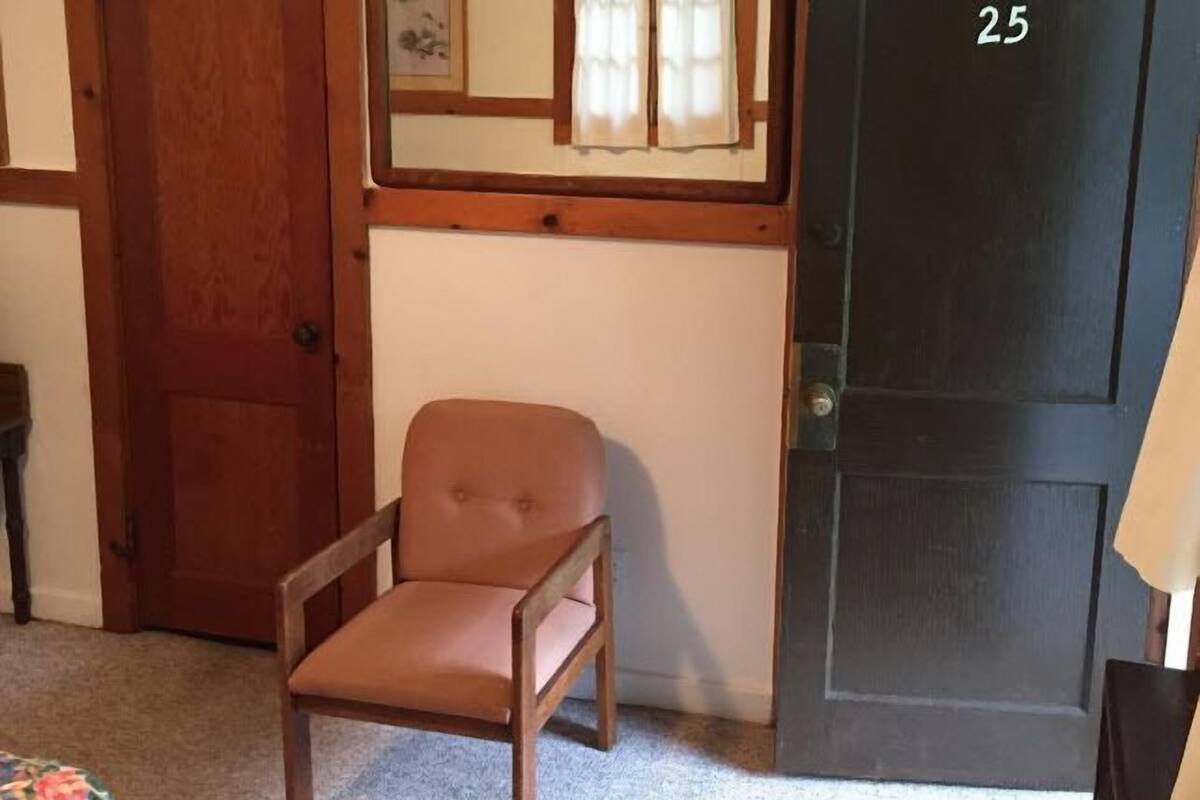 A room with a wooden door marked with the number 25, a brown chair, a small table, and a wall-mounted mirror.