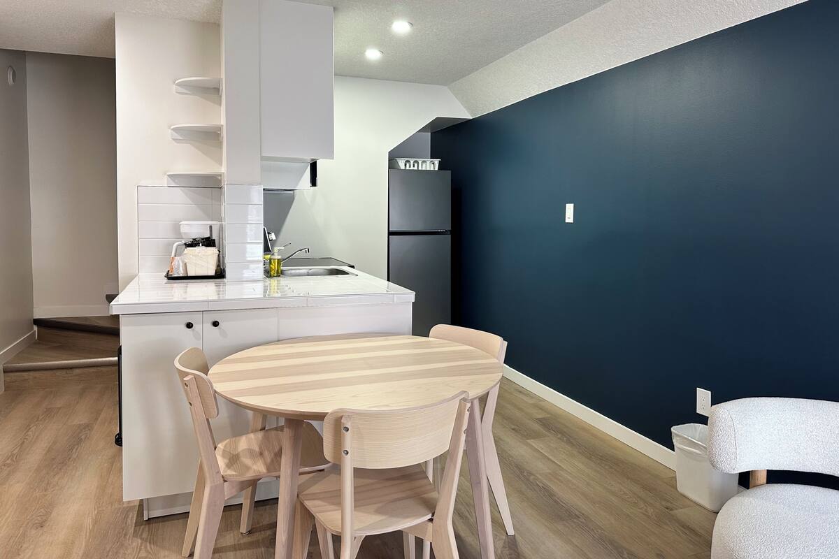 A compact kitchen with a white countertop, a sink, and a built-in refrigerator. There is a round wooden dining table with chairs. The walls are painted in two colors, one dark and one light.