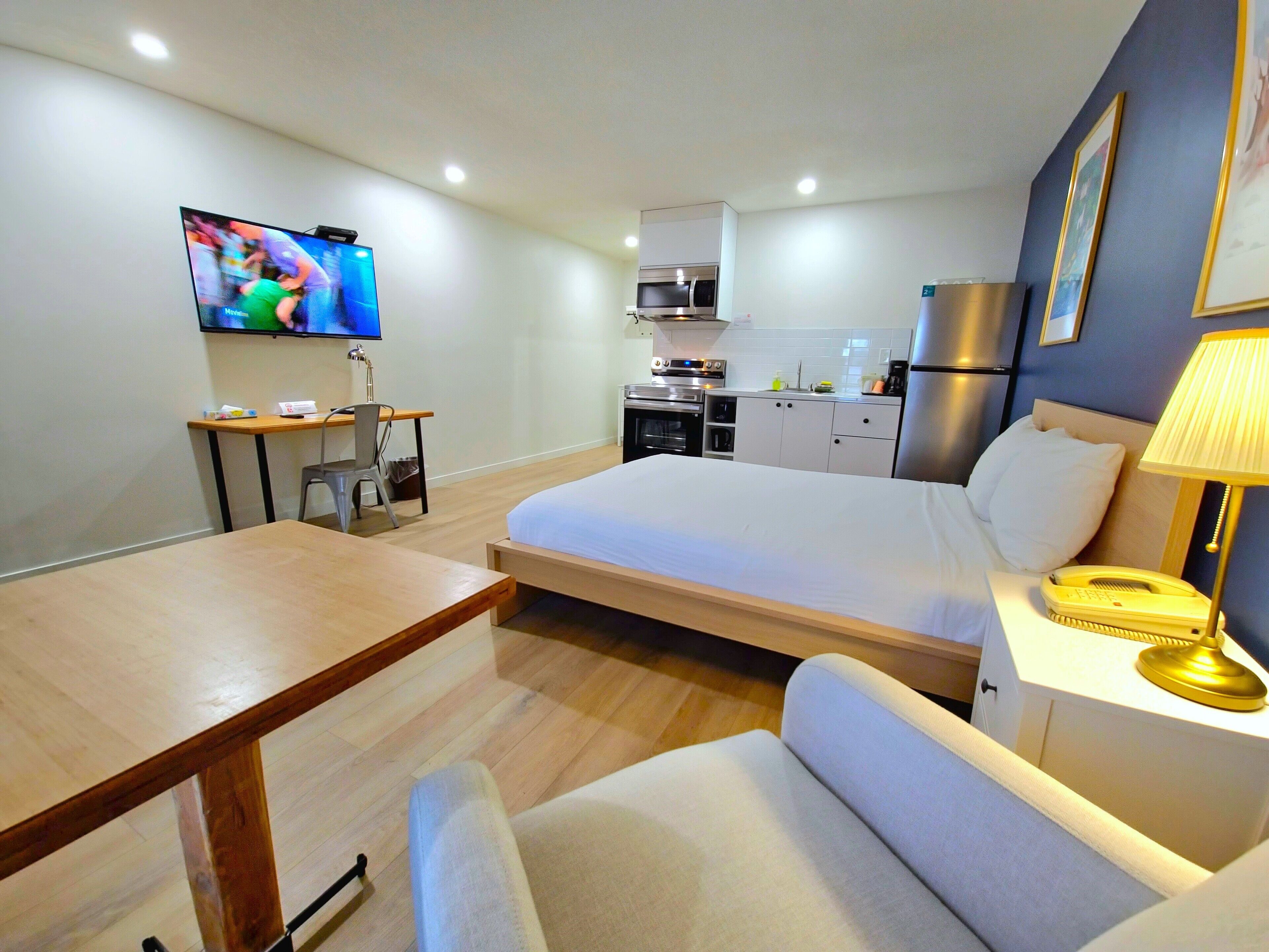 A modern hotel room with a bed, a desk, a chair, a TV, a kitchen area, and a lamp.