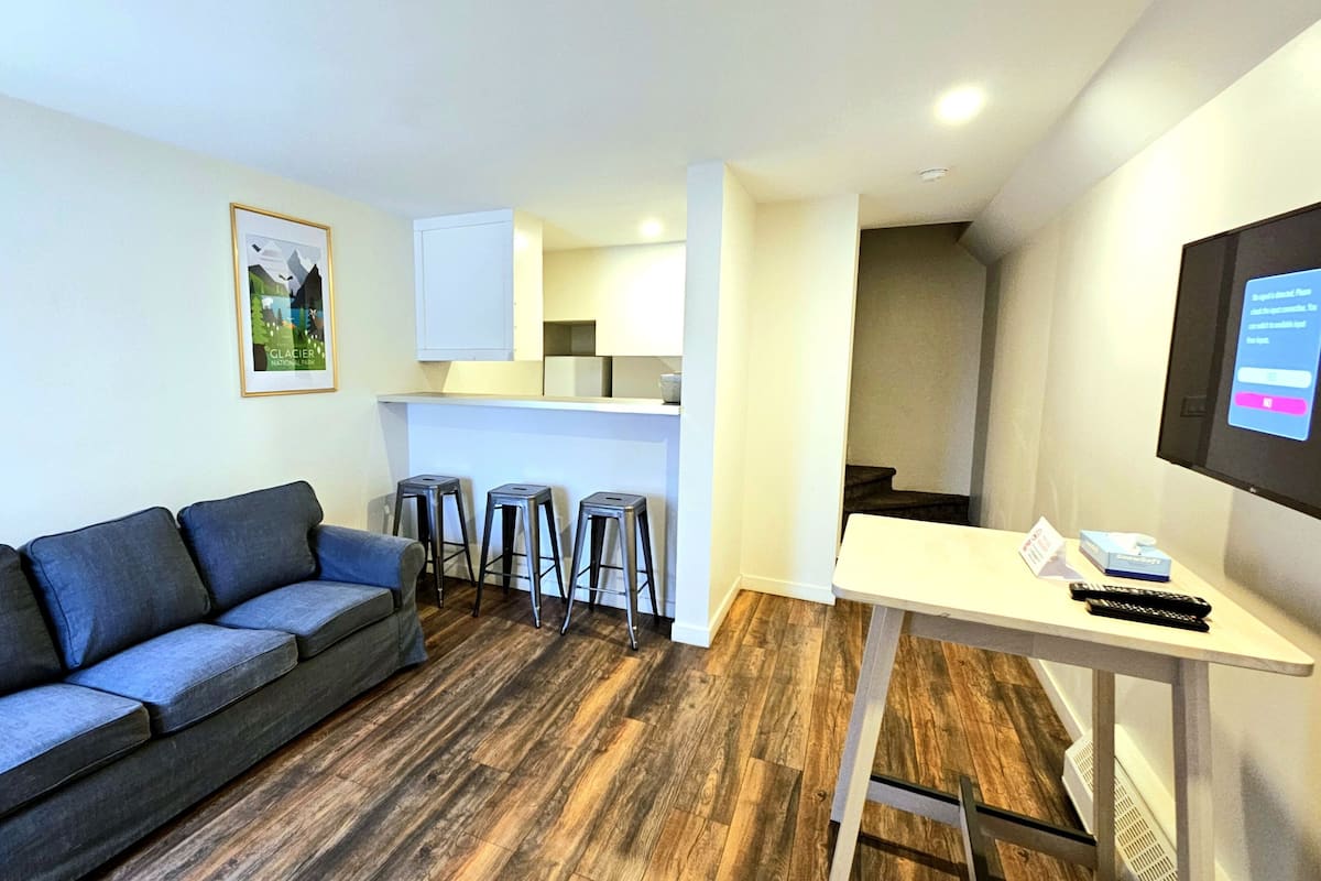 A compact living space with a grey sofa, three metal stools, a white table with a remote control, and a framed picture on the wall.