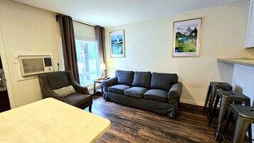 A living room with a sofa, armchair, and a table. There are two framed pictures on the wall, a window with curtains, and a radiator.