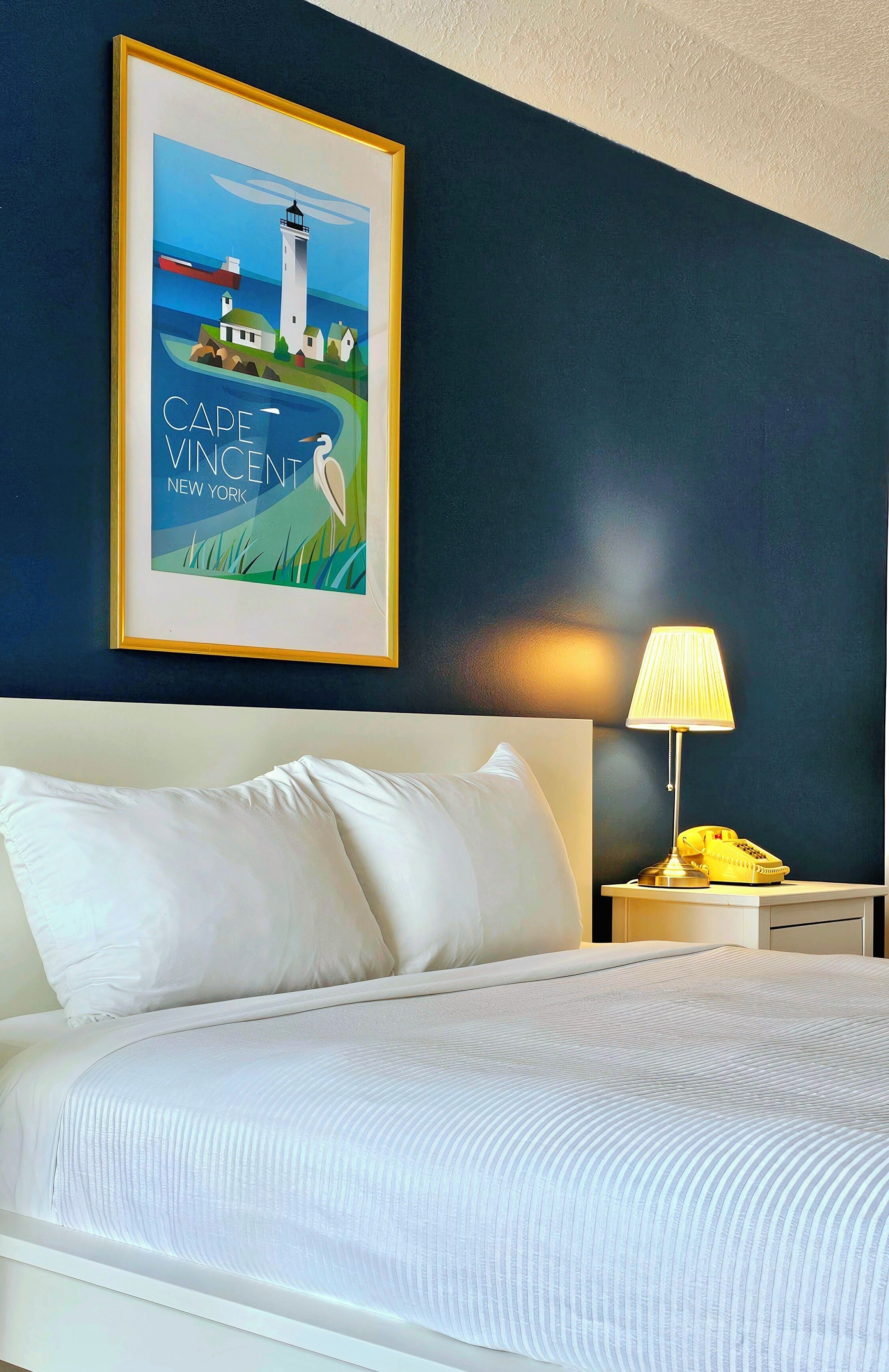 A neatly made bed with white linens, a framed picture of Cape Vincent, New York, on the wall, and a bedside lamp with a telephone.