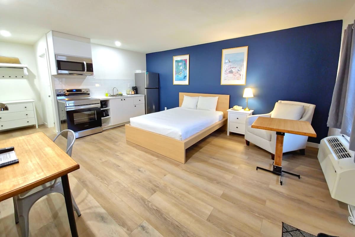 A hotel room with a bed, bedside table, chair, desk, kitchenette, and artwork on the wall.