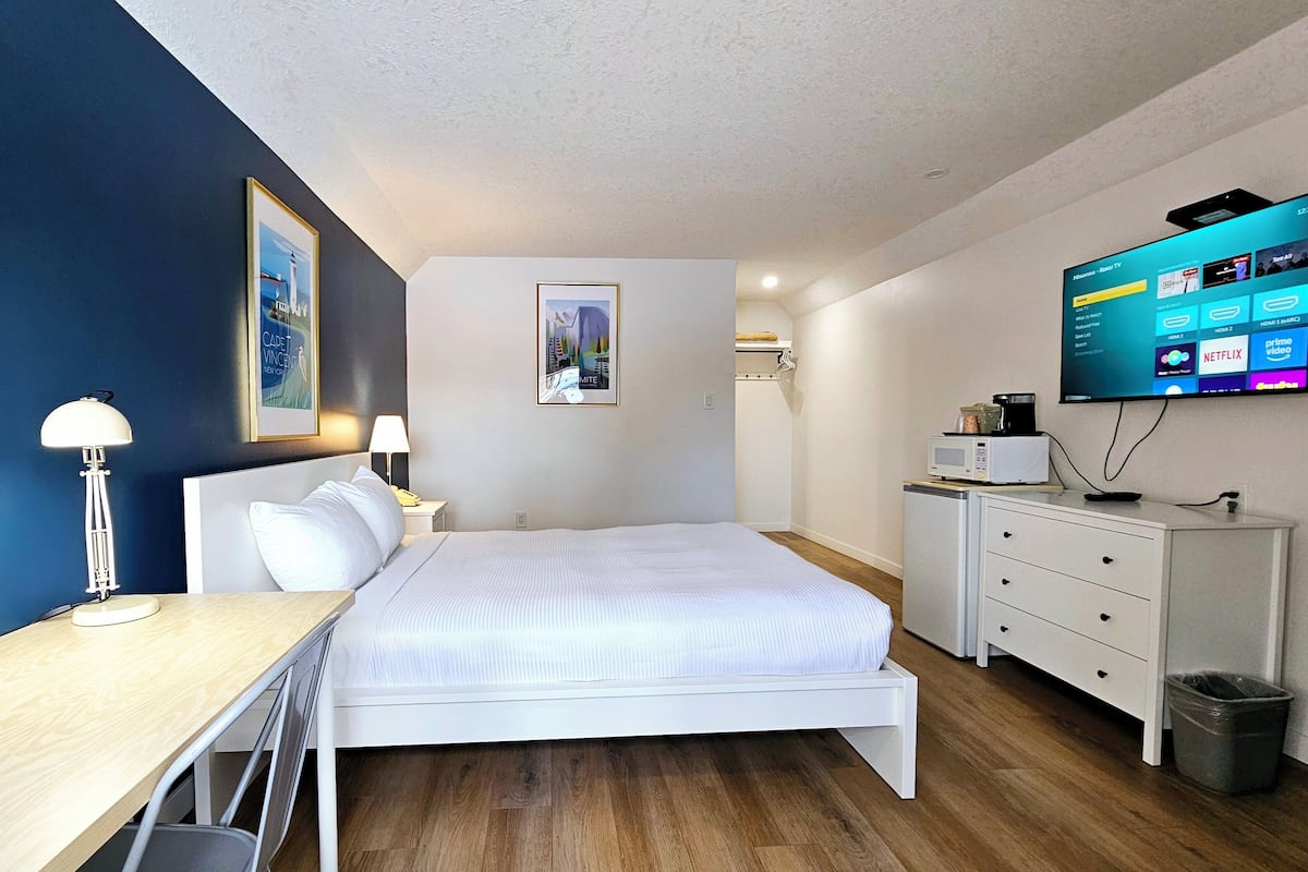 A hotel room with a bed, desk, lamp, TV, and a trash bin.