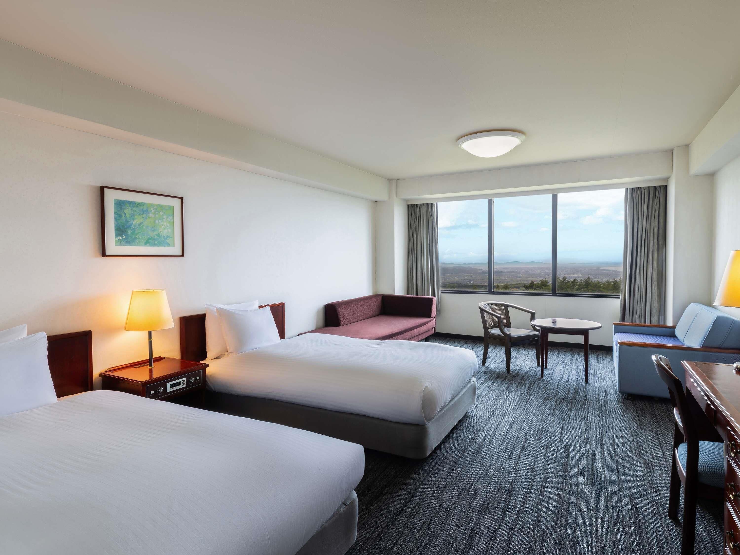 Quarto de hotel com duas camas, uma escrivaninha, uma cadeira, um sofá e uma janela grande com vista.