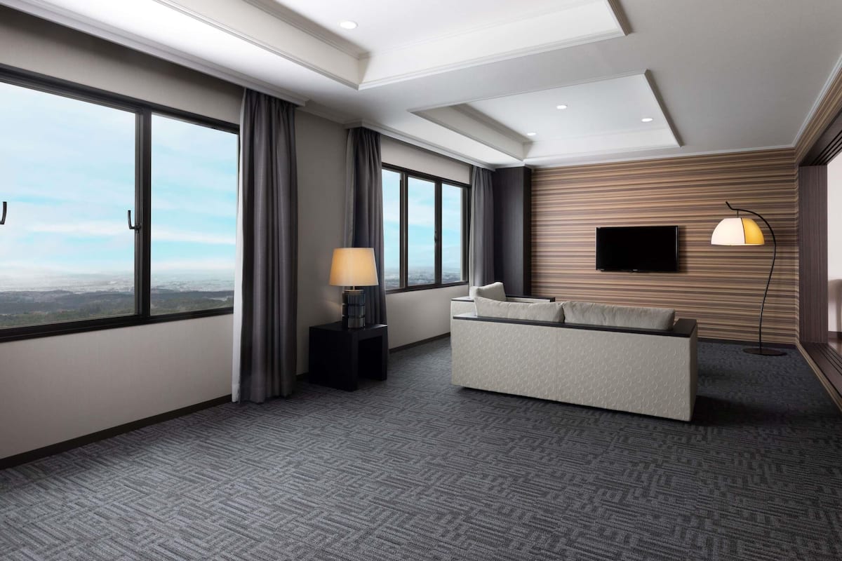 Una habitación de hotel moderna con ventanas amplias, un sofá, un televisor de pantalla plana y una lámpara.