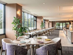 Una larga mesa de comedor con sillas, ventanas amplias y plantas en macetas.