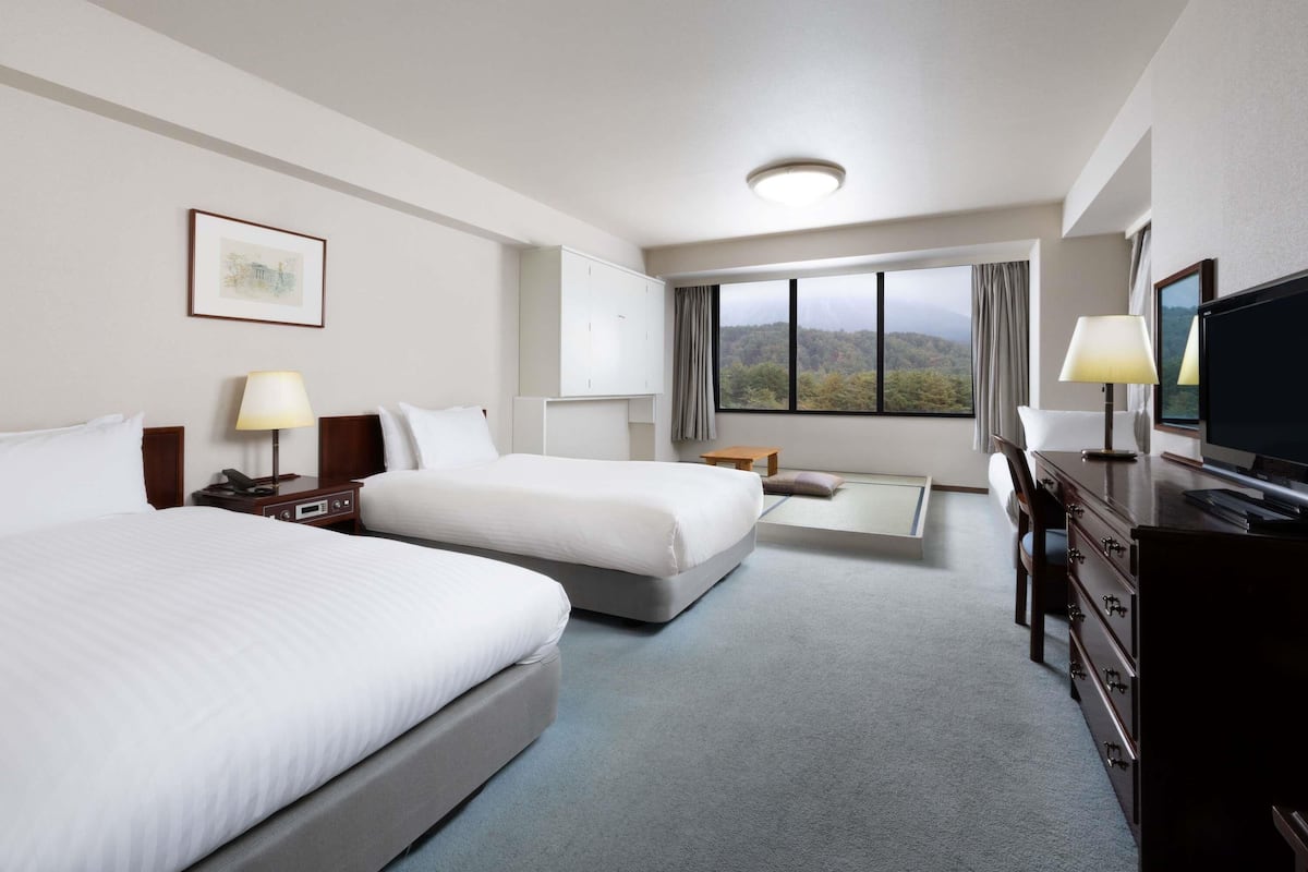 A hotel room with two beds, a desk, a television, and a large window with a mountain view.