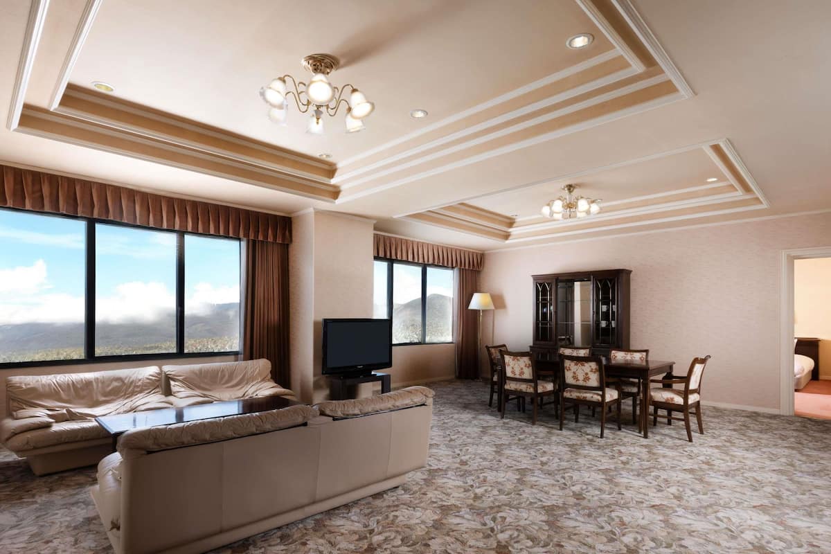 A hotel room with a large window, a sofa, a TV, a dining table with chairs, and a chandelier.