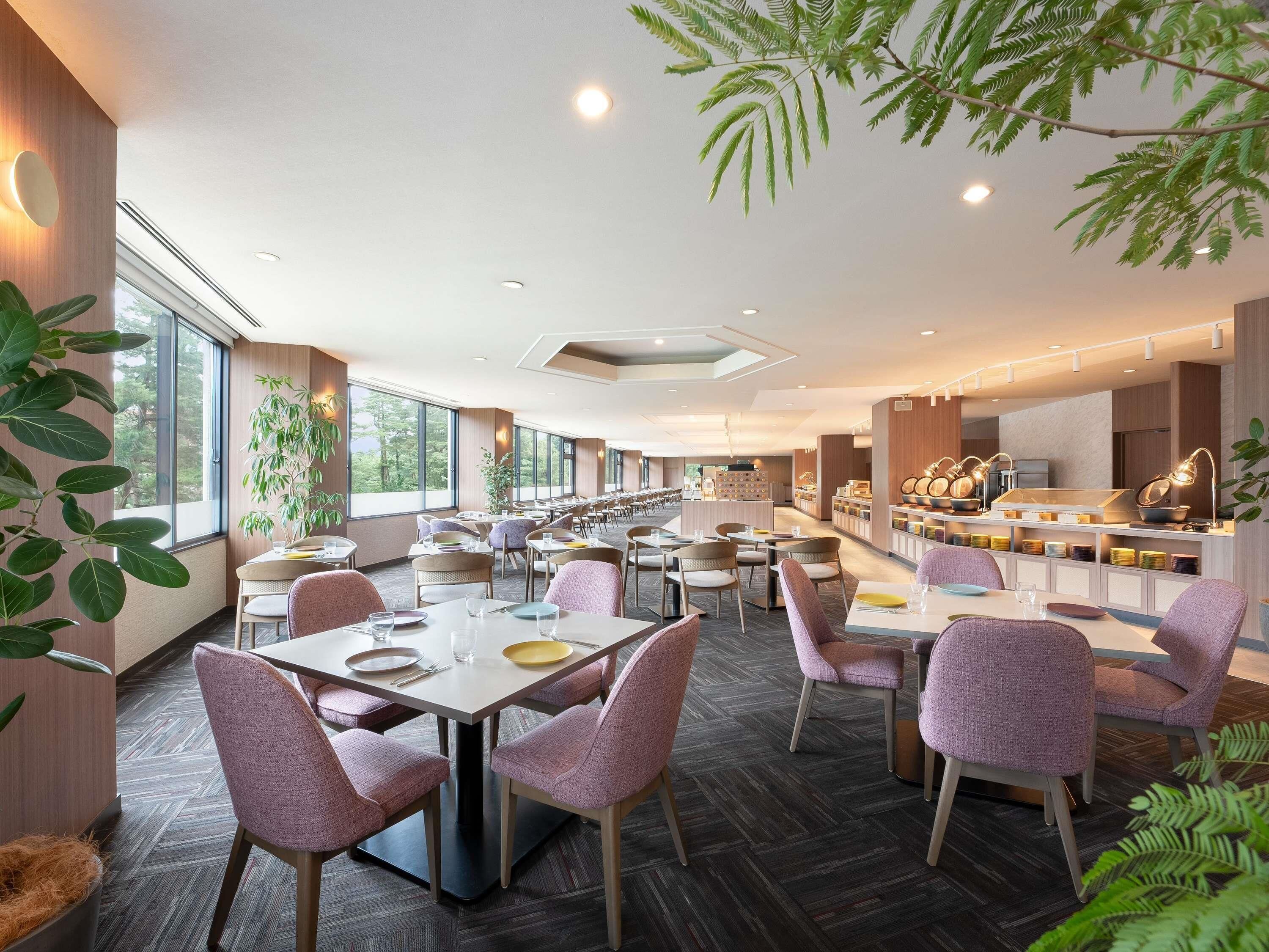 A spacious restaurant with multiple dining tables and chairs, large windows, and indoor plants.