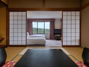 A traditional Japanese-style room with tatami flooring, a bed, a television, and a view of a forested landscape.