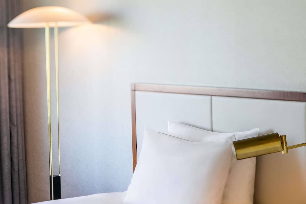A neatly made bed with white linens and pillows, a gold-colored headboard, and a floor lamp with a white shade.