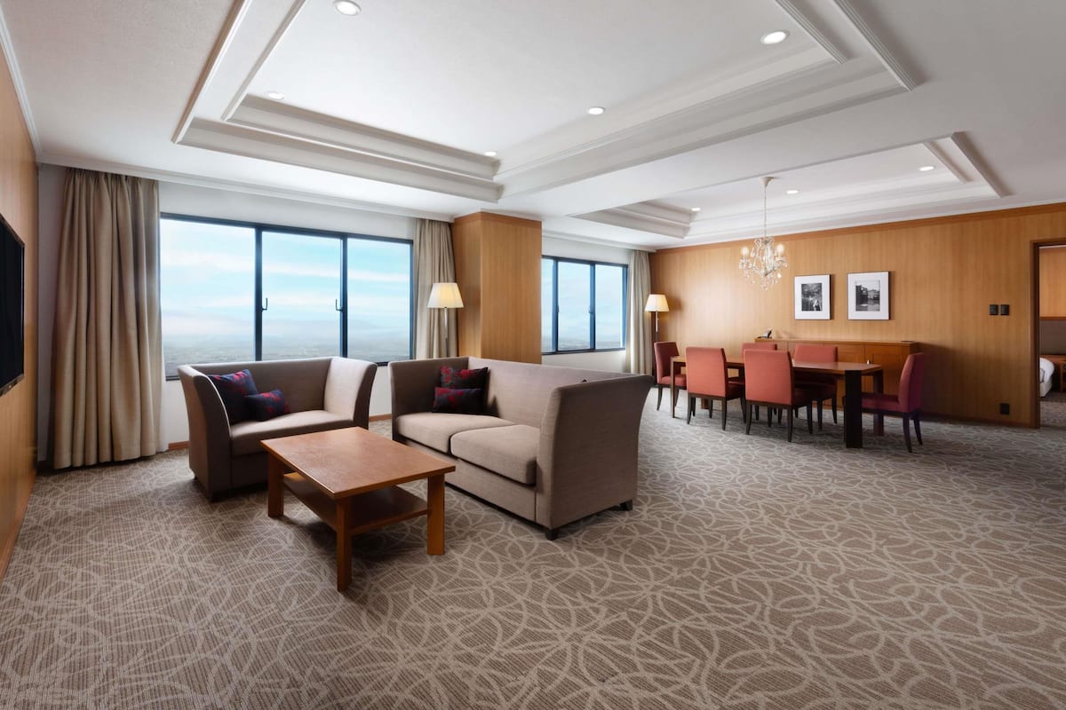 A hotel room with a sofa, a small table, a dining area with chairs, and a large window offering a view.