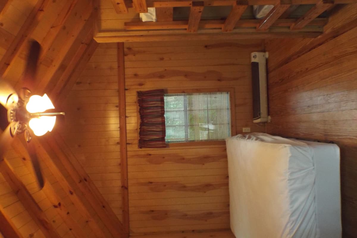 A wooden cabin interior with a bed, a window with blinds, and a ceiling light fixture.