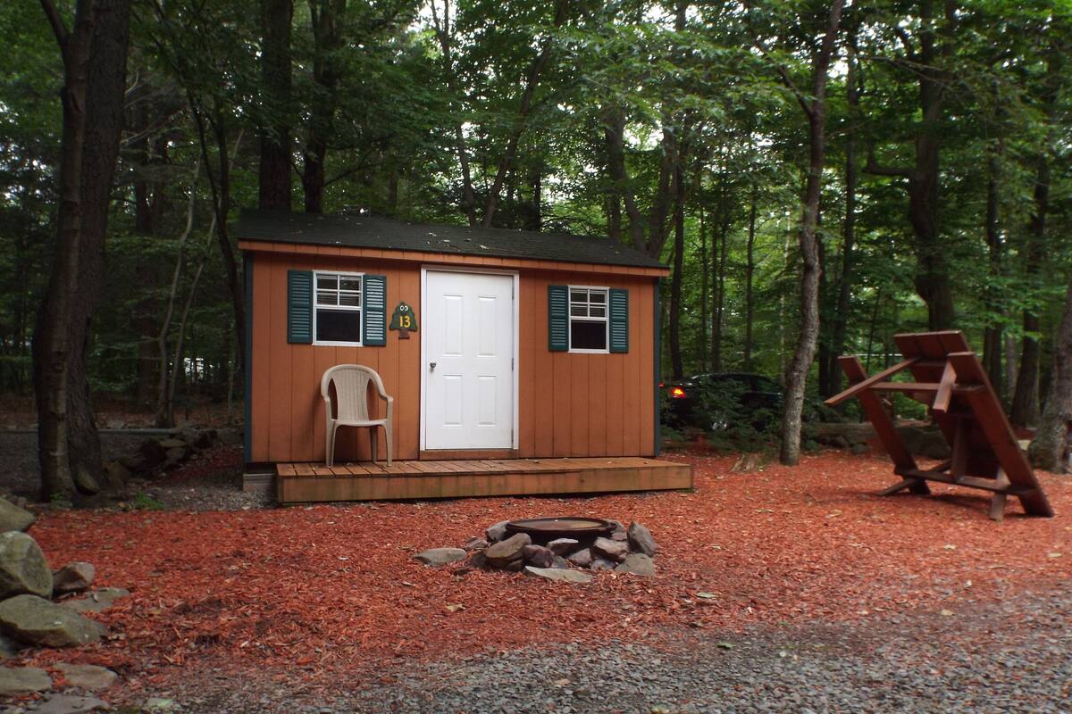 A small wooden cabin with a white door, two windows with shutters, a chair outside, and a fire pit in front.