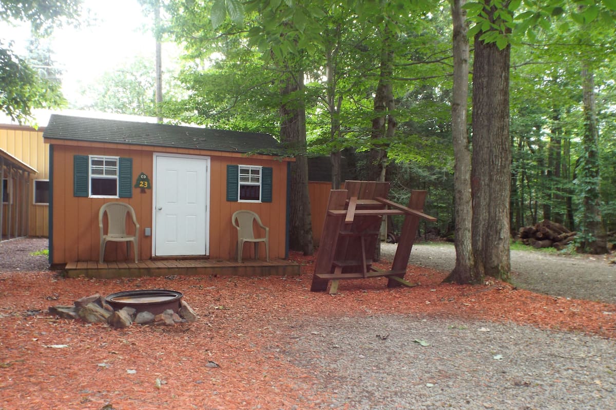 A small cabin with a white door, two windows with green shutters, and a porch area with two chairs.