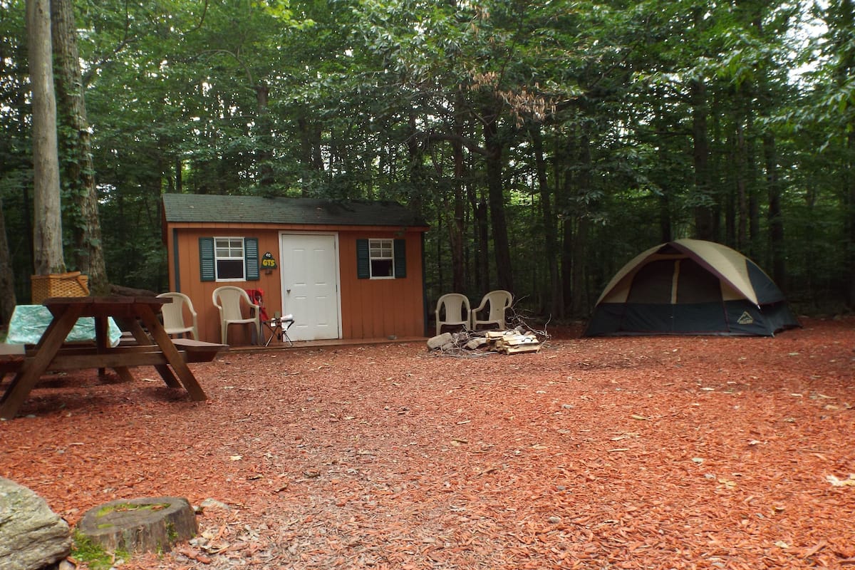 A campsite with a small cabin, a tent, picnic tables, and a fire pit.