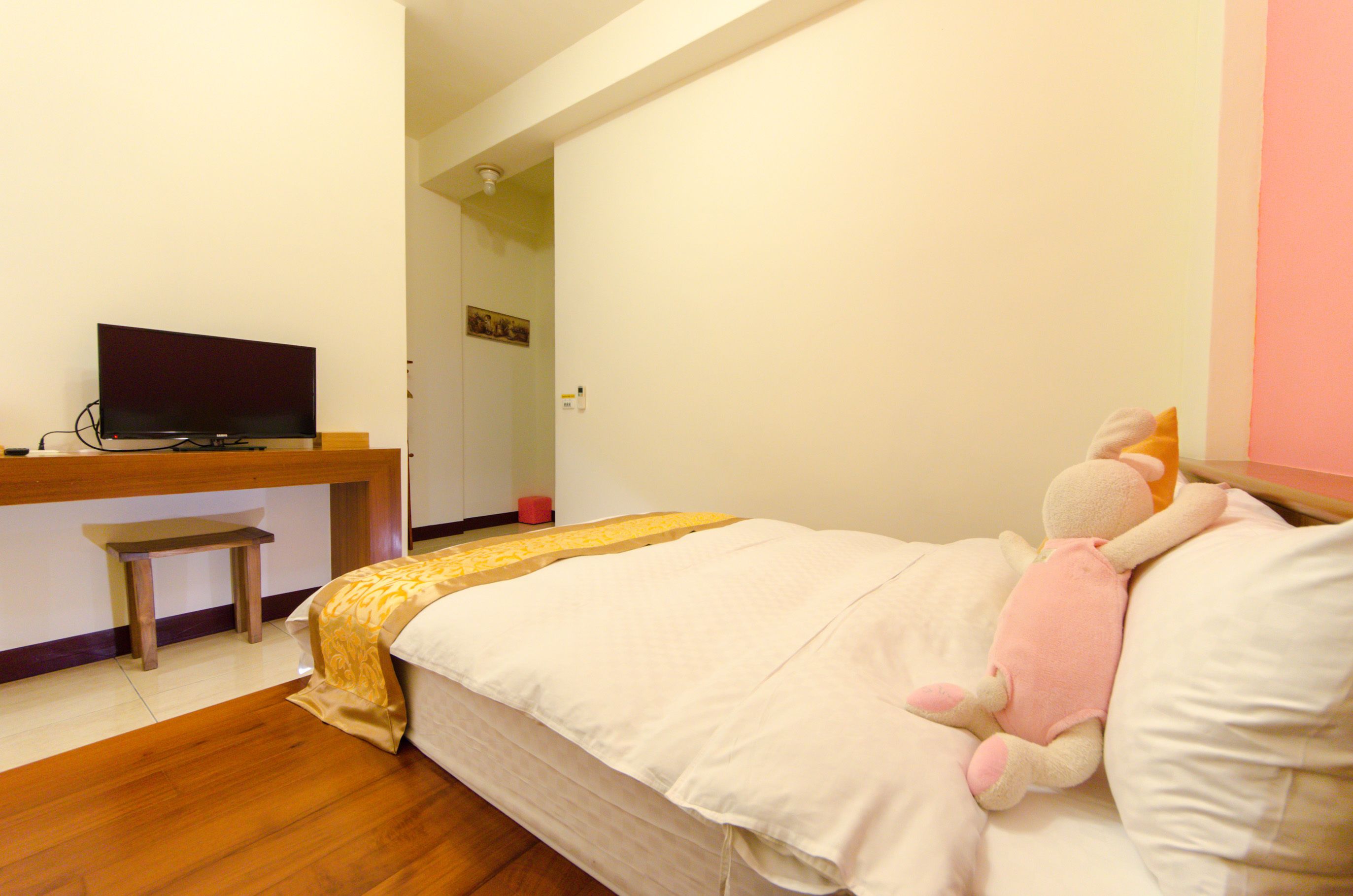 Un dormitorio con una cama, un escritorio con televisor, un pequeño banco y un peluche sobre la cama.
