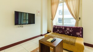 Un salon avec un téléviseur à écran plat fixé au mur, un canapé agrémenté d’un plaid à motifs et une petite table en bois sur laquelle repose une télécommande.