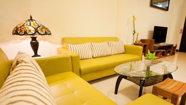 Un canapé jaune, une table basse en verre et un petit tabouret en bois dans un salon agrémenté d’une lampe et d’une plante.