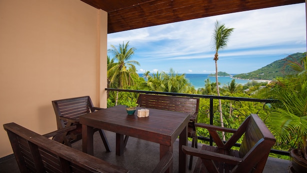 A wooden table and chairs on a balcony with a view of the ocean and palm trees.