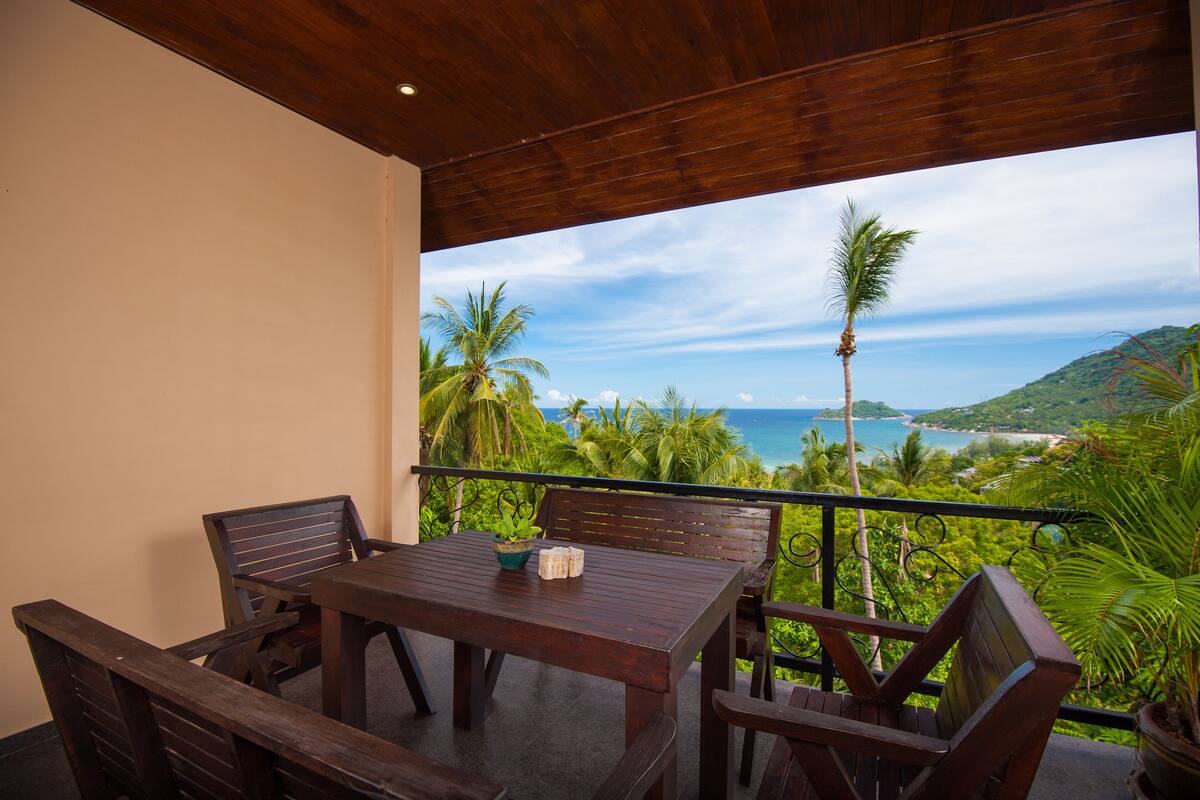 A wooden table and chairs on a balcony with a view of the ocean and palm trees.