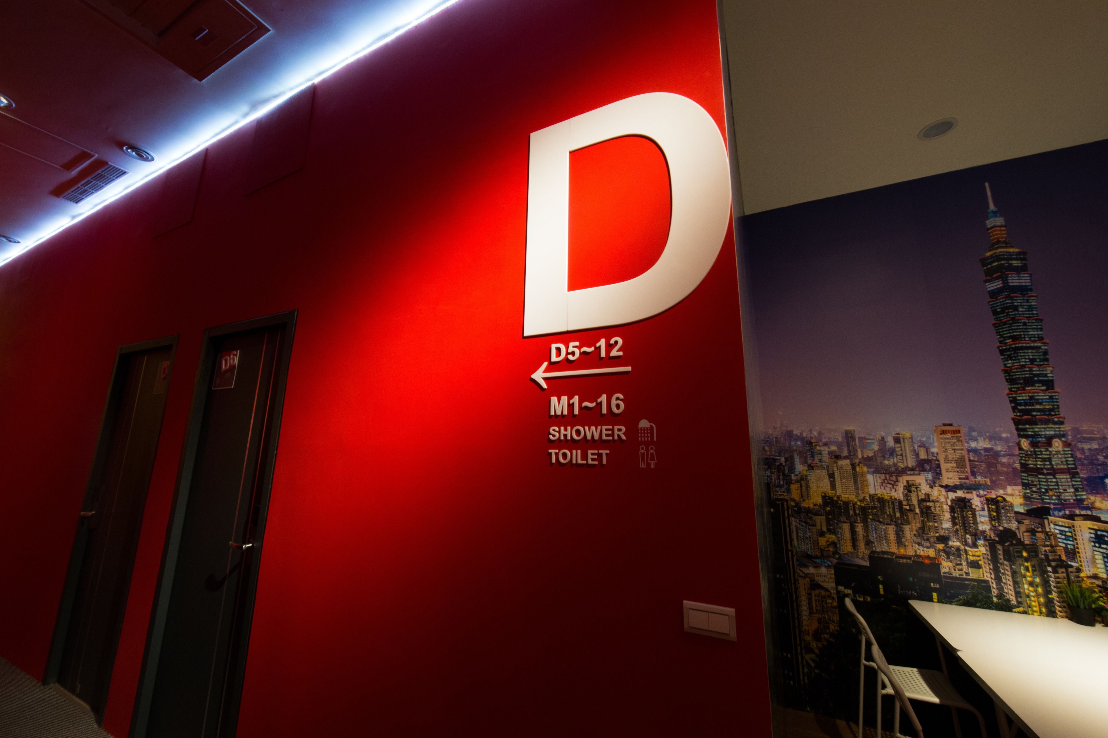 A hotel corridor with a red wall, a large 'D' sign indicating room D5-12, and a poster of a cityscape with a tall building.