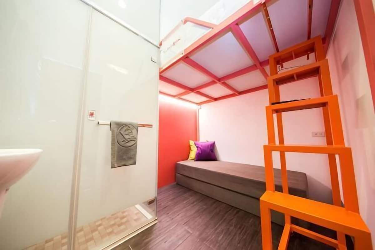 A compact room with a glass shower enclosure, a small bed with a pillow, and a wooden ladder.