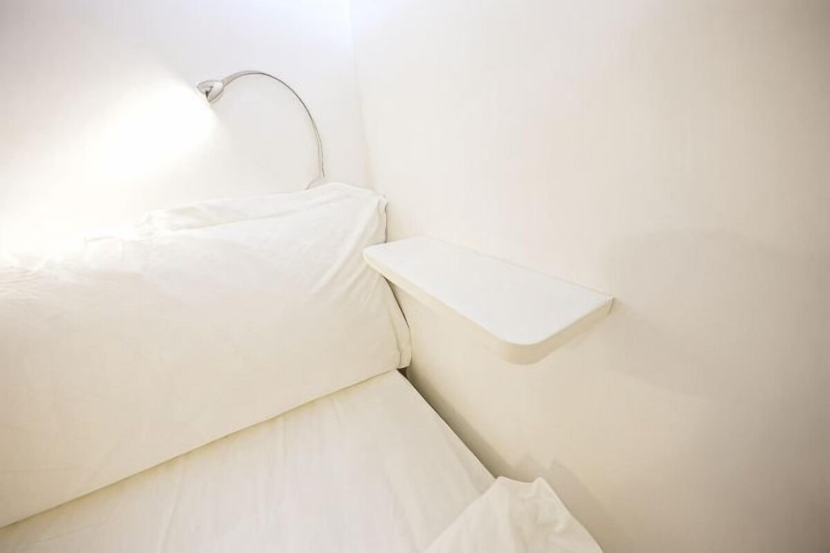 A white bed with a headboard, a bedside table, and a wall-mounted light fixture.
