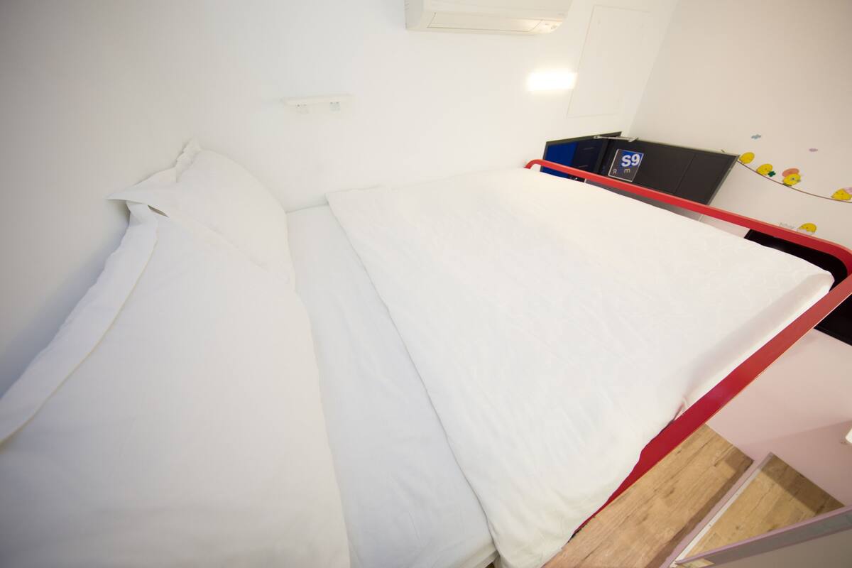 A single bed with white bedding, a red frame, and a small bedside table with a blue object on it.