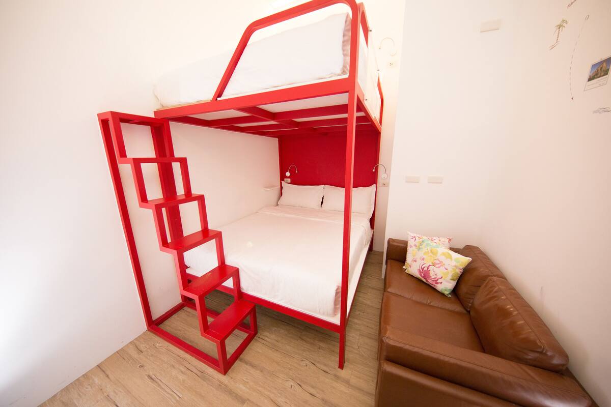 A bunk bed with a red frame and white bedding, a brown leather sofa, and a small wall-mounted shelf.