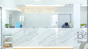 A reception desk with a marble wall, a reception sign, a cart with amenities, and a person at the desk. - Bed by Tha-Pra Hotel and Apartment (Bangkok)
