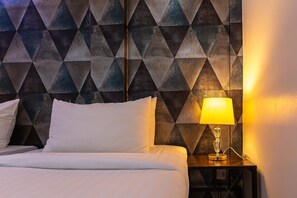 Una cama bien hecha con sábanas y almohadas blancas, una mesita de noche con lámpara, y una pared con un diseño geométrico.
