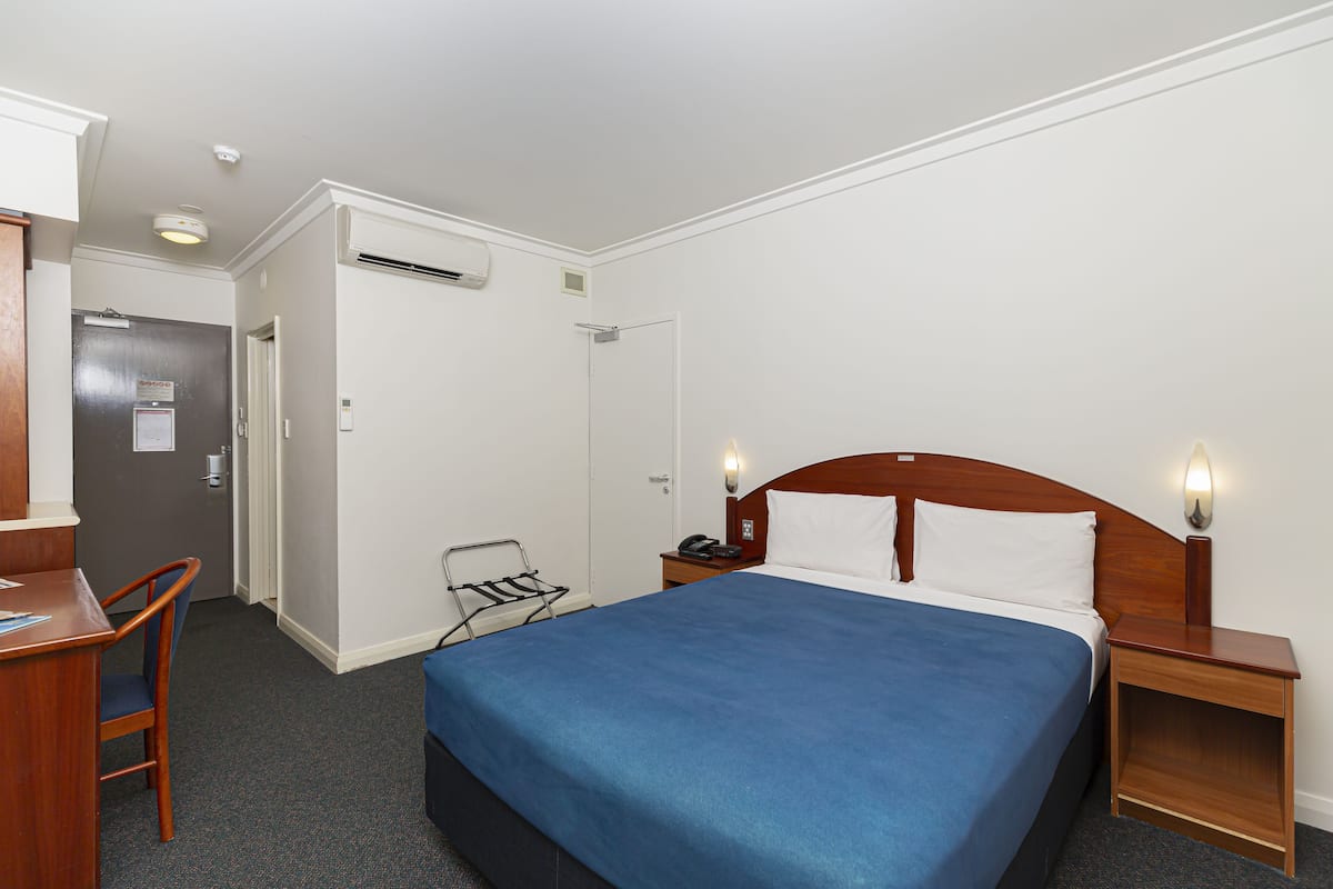 Una habitación de hotel con una cama individual, un escritorio, una silla, una mesita de noche con una lámpara y una puerta que da a otra habitación.