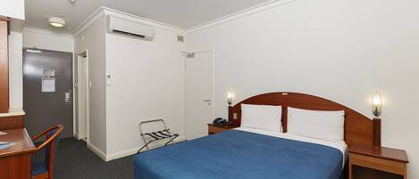 A hotel room with a single bed, a desk, a chair, a nightstand with a lamp, and a door leading to another room.