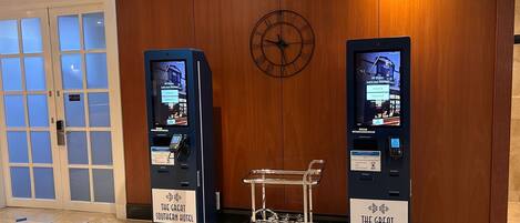 A hotel lobby with two self-service kiosks, a digital clock, and a luggage cart.