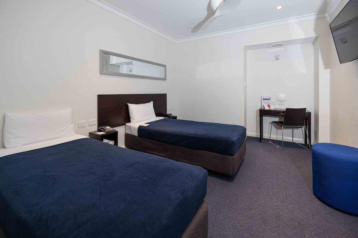 Habitación de hotel con dos camas individuales, un escritorio con silla, televisión montada en la pared y un espejo sobre la cama.