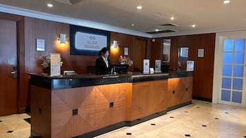 A hotel reception desk with a wooden panel design, a digital screen displaying the hotel name, and a receptionist.