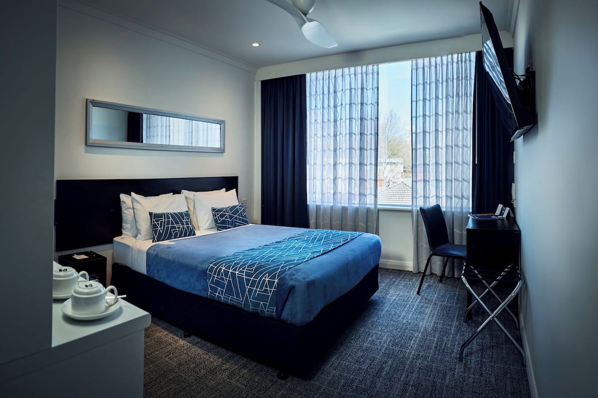 Habitación de hotel con una cama grande, un escritorio con silla, un televisor montado en la pared y un ventanal con cortinas.
