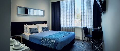 A hotel room with a large bed, a desk with a chair, a TV mounted on the wall, and a window with curtains.