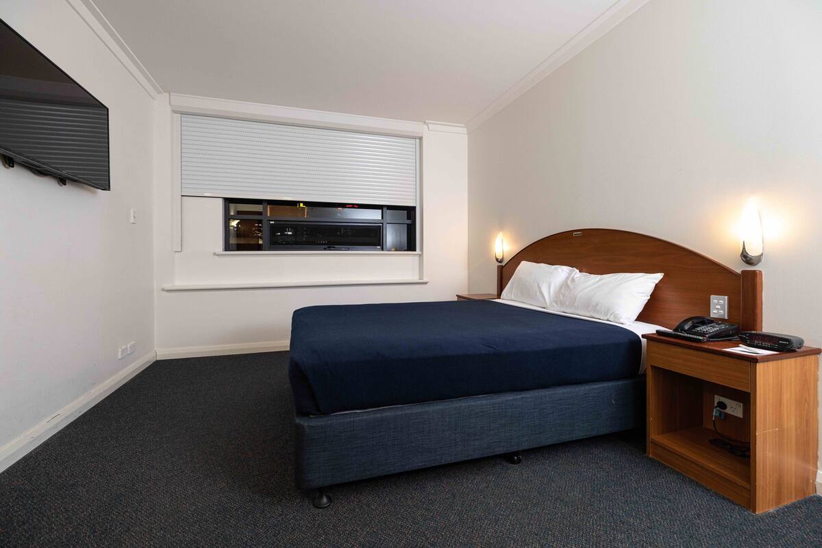 Habitación de hotel con una cama, una mesita de noche con lámpara, un televisor montado sobre la cama y una ventana con persianas.