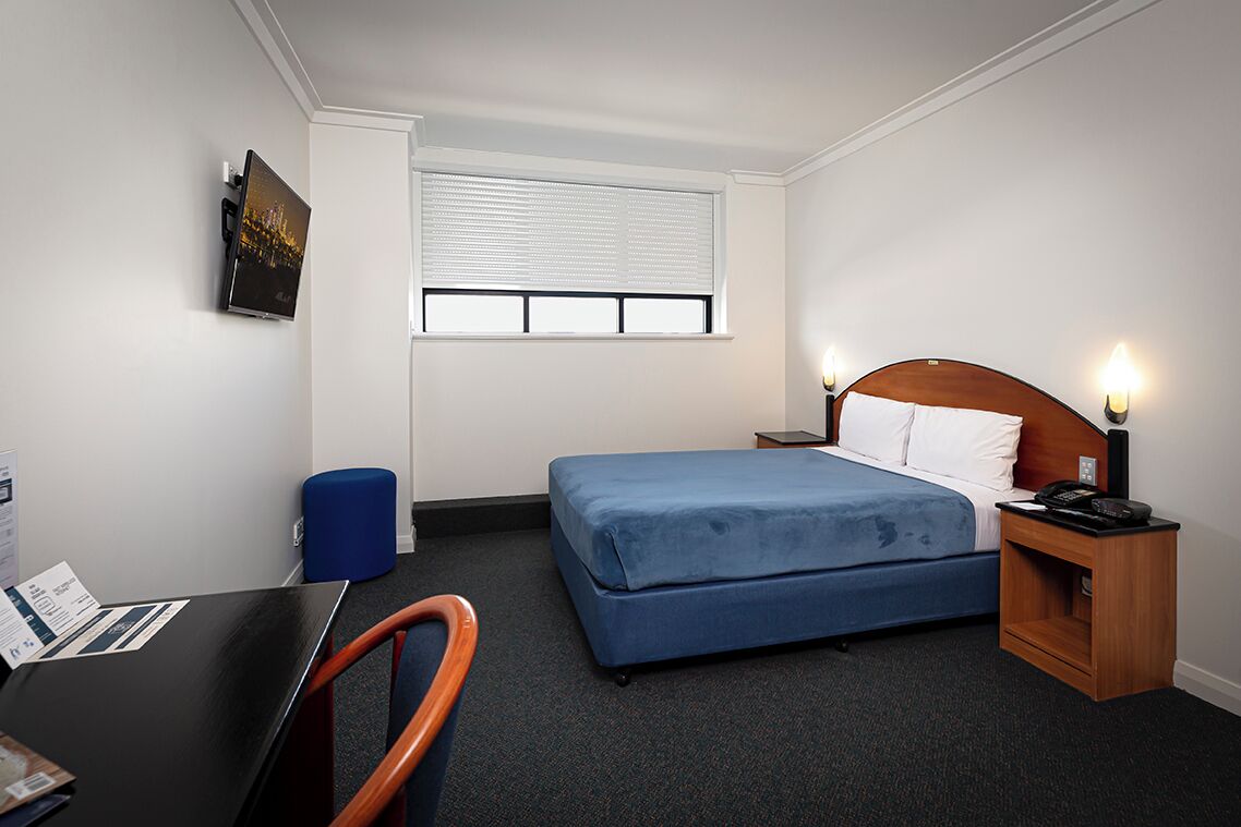 A hotel room with a bed, bedside table, lamp, chair, desk, and a window with blinds.