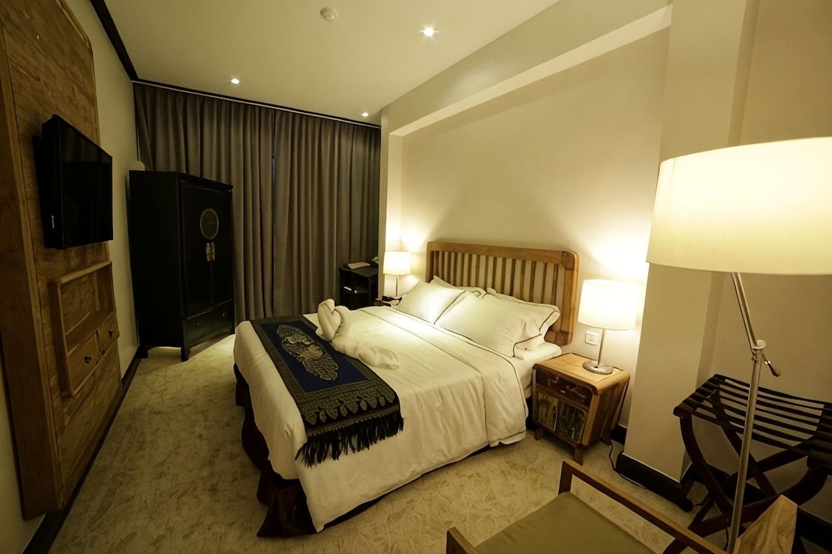 Habitación de hotel con cama, lámparas de noche, cabecera de madera, mesita de noche, televisor y armario.