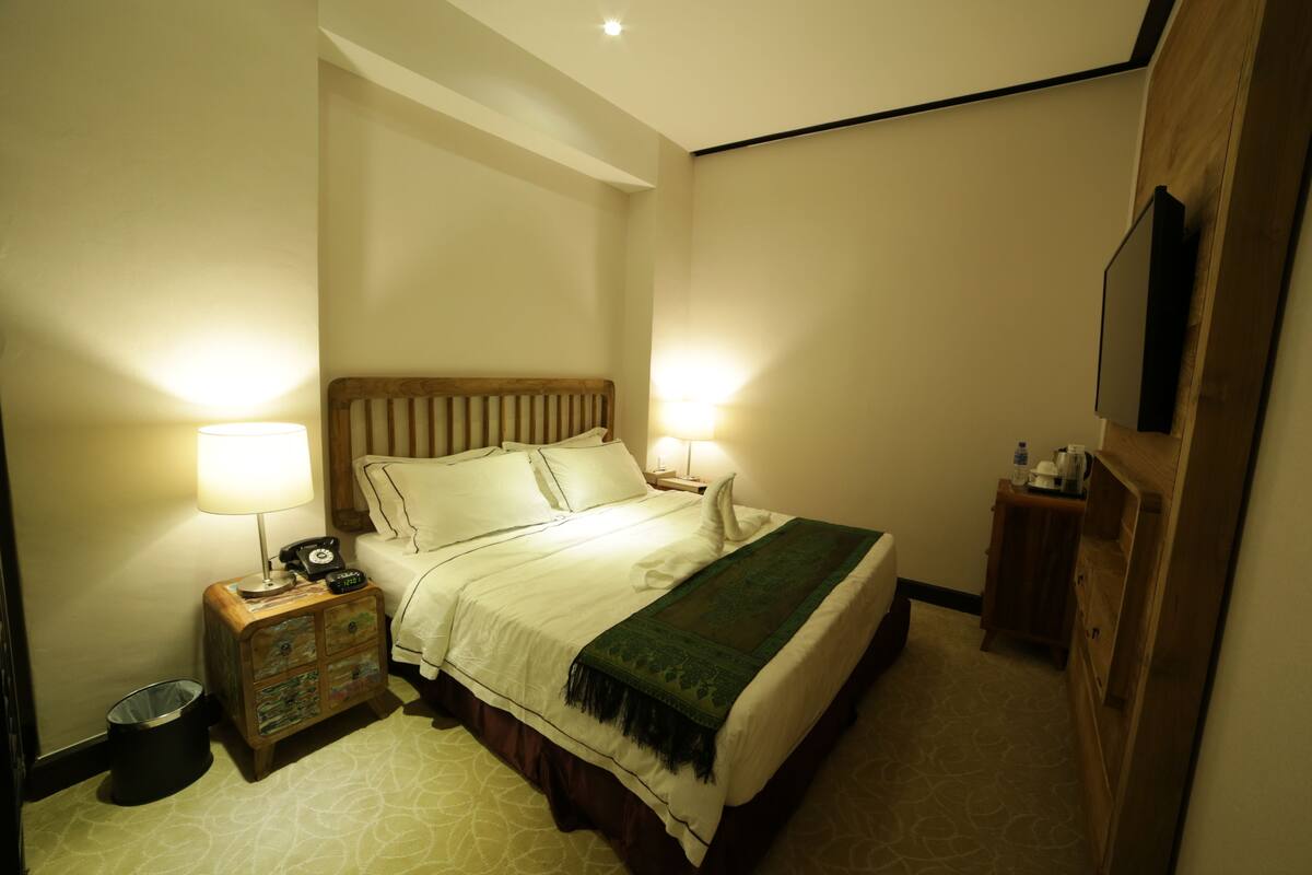 Une chambre d’hôtel comprenant un lit, des lampes de chevet, une table de chevet en bois, une poubelle et une commode en bois.