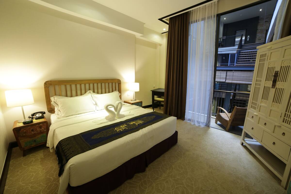 Une chambre d’hôtel avec un grand lit, une table de chevet avec une lampe, une commode et une vue sur le balcon extérieur.