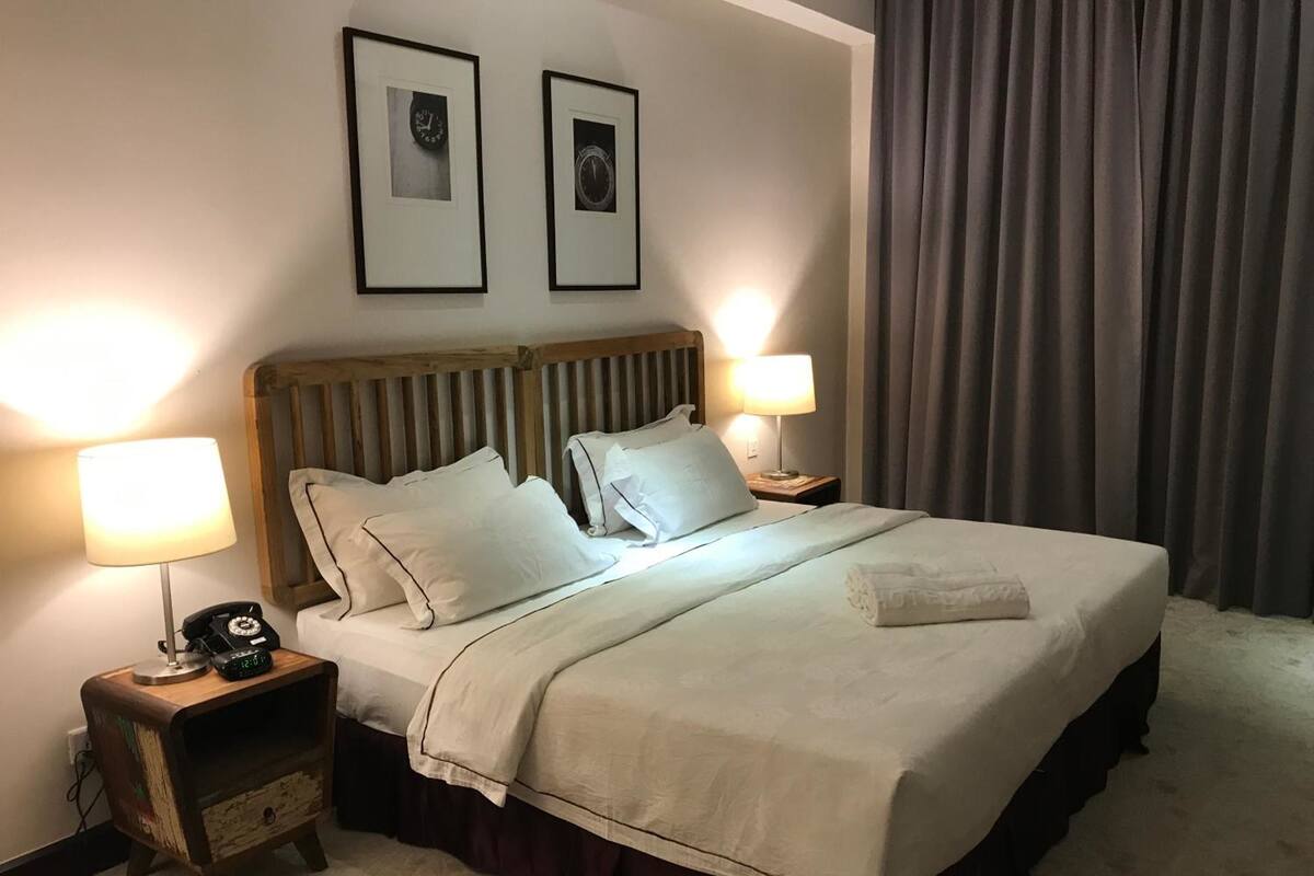 Un lit bien fait, avec une couette blanche et des oreillers, deux lampes de chevet, une table de chevet en bois avec un téléphone, et un tableau encadré au-dessus du lit.