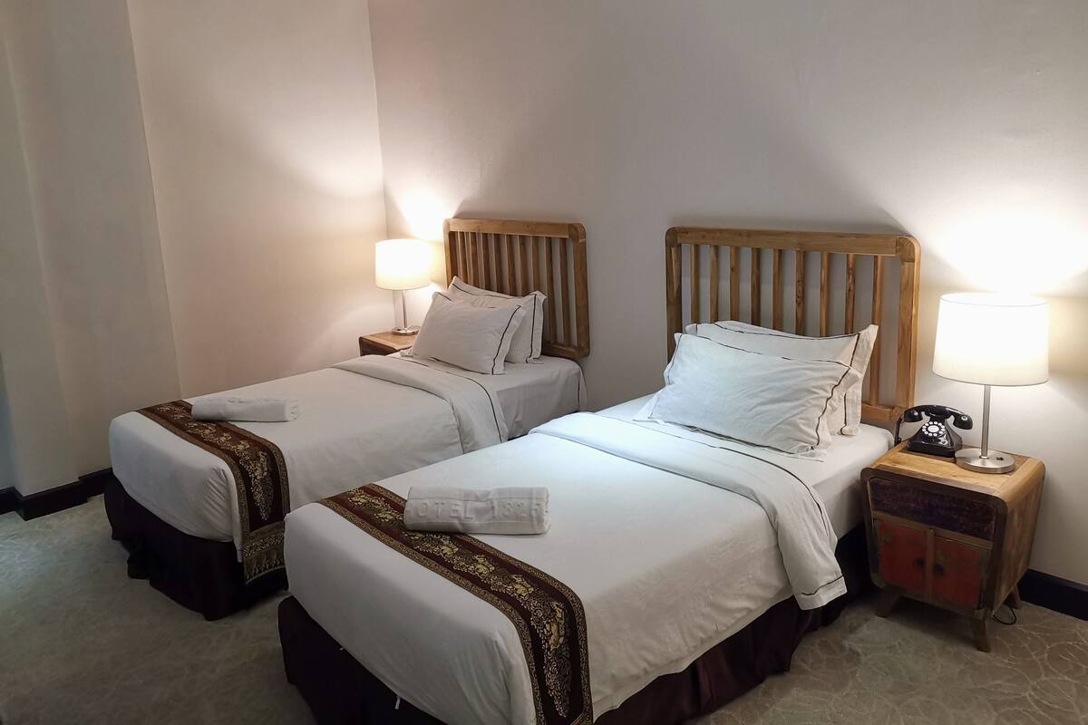 Une chambre d’hôtel avec deux lits, chacun doté d’une tête de lit en bois, de draps blancs et d’une petite table de chevet avec une lampe.