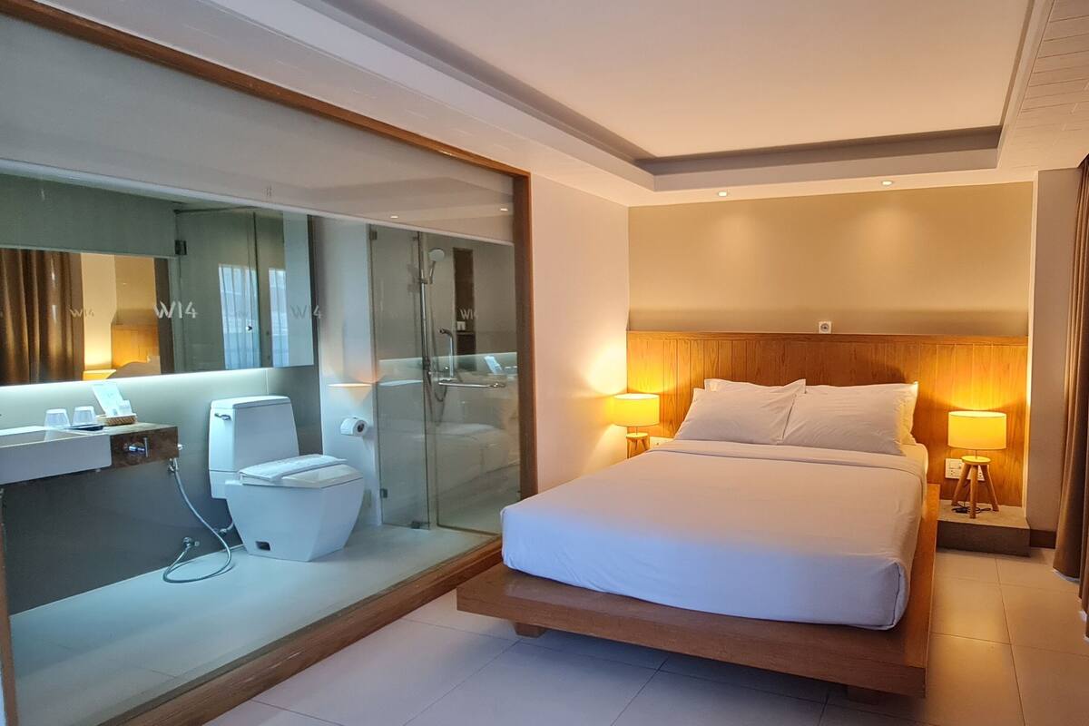 A modern hotel room with a large bed, bedside lamps, a glass shower enclosure, and a wooden headboard.