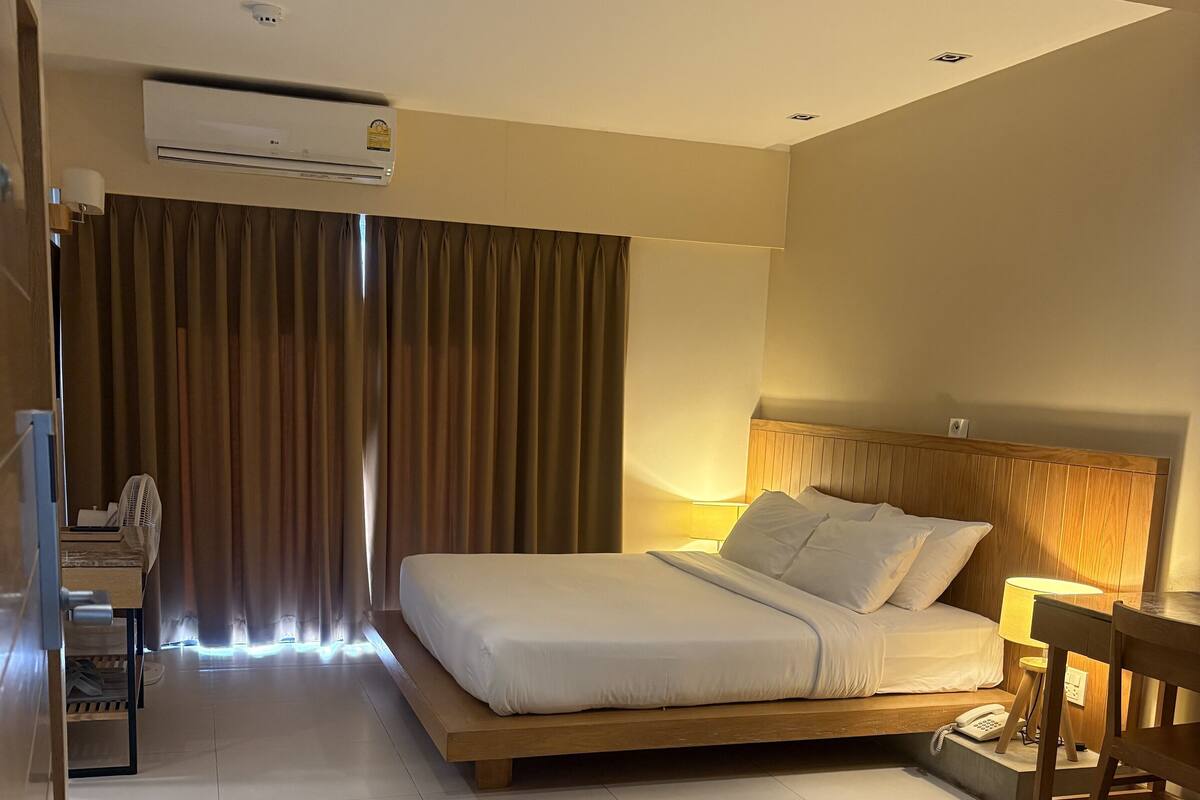 A modern hotel room with a large bed, a desk, a chair, an air conditioning unit, and a fan.