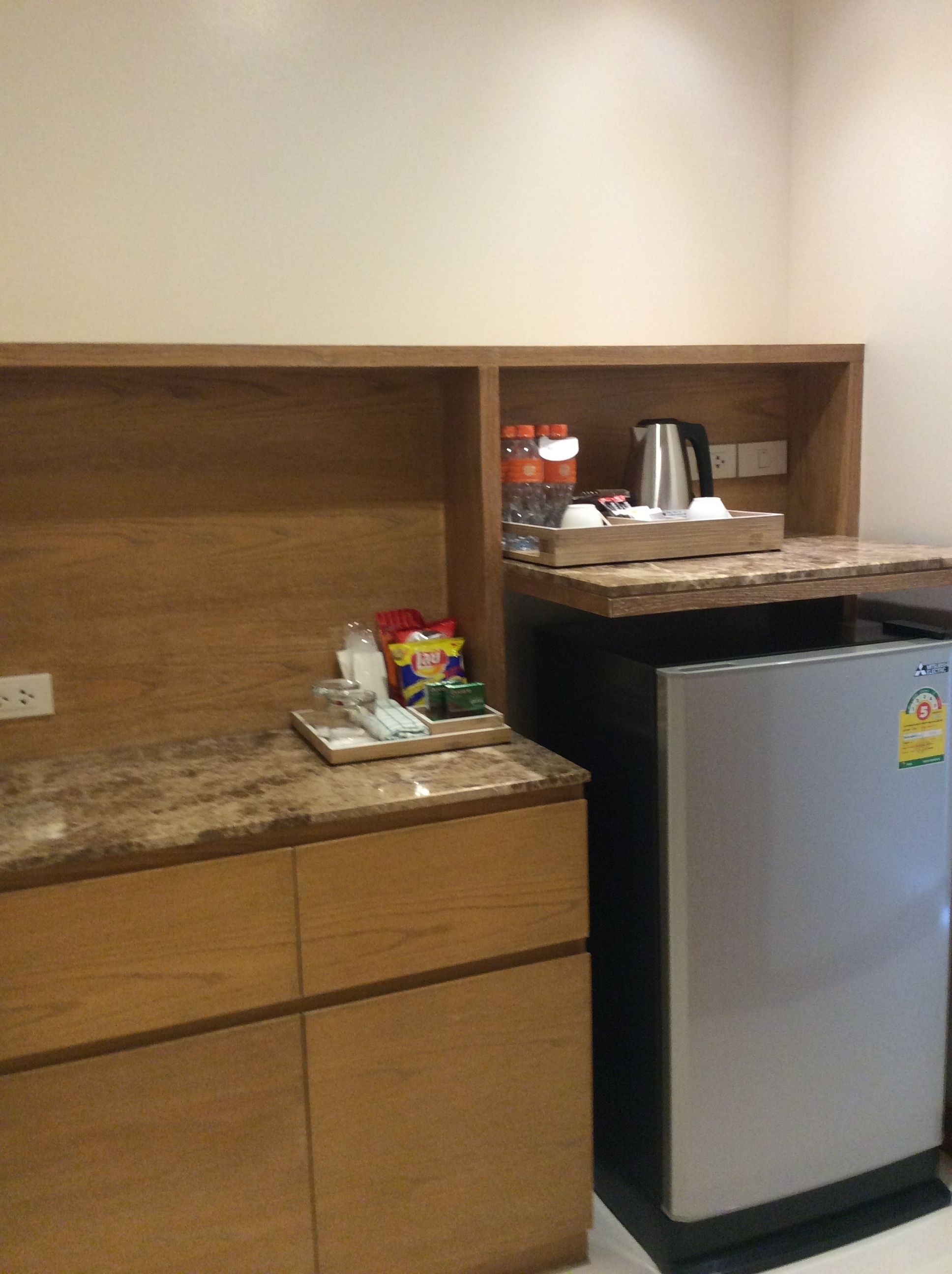 A compact kitchen with wooden cabinetry, a stainless steel refrigerator, and a countertop with a kettle and a coffee maker.